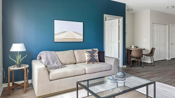 Een woonkamer met een beige bank, een glazen salontafel en een blauwe accentmuur.