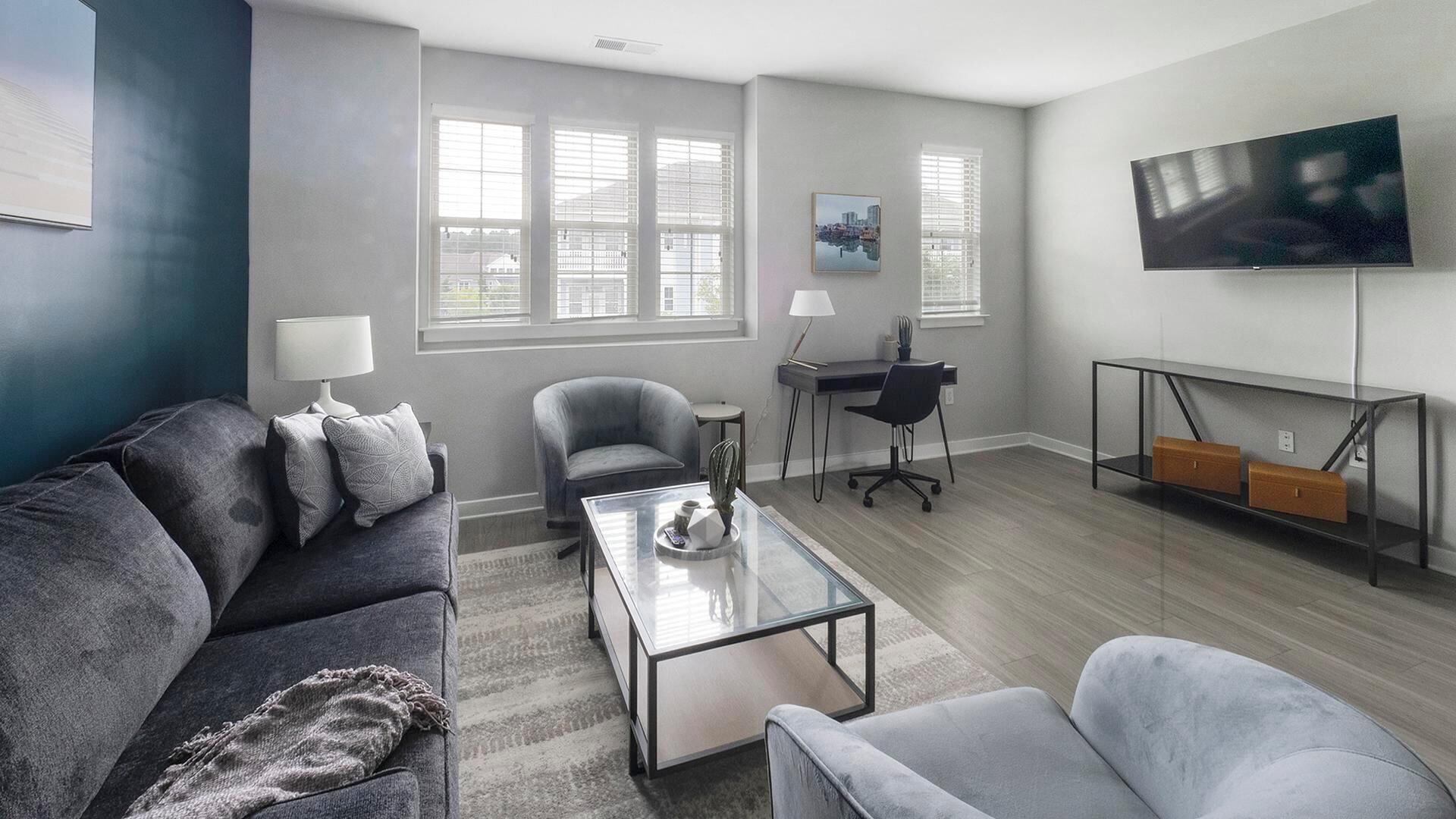 A modern living room with a grey sofa, a glass coffee table, and a grey armchair. There is a desk with a chair and a TV mounted on the wall.