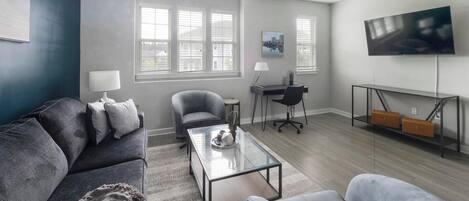 A modern living room with a grey sofa, a glass coffee table, and a grey armchair. There is a desk with a chair and a TV mounted on the wall.