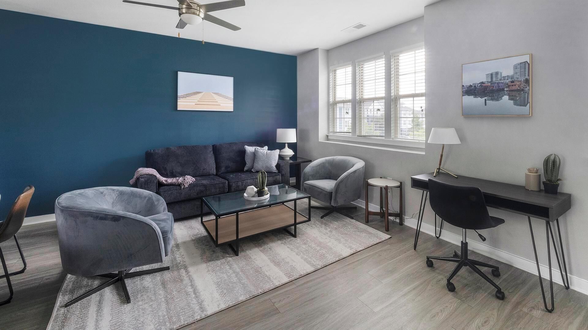 Una sala de estar moderna con una pared de acento azul oscuro, un ventilador de techo, un sofá, dos sillones, una mesa de centro, un escritorio con una silla y una ventana con persianas.