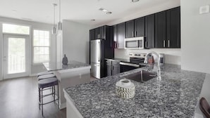 A modern kitchen with dark cabinets, stainless steel appliances, a granite countertop, and a bar stool. - Landing | Incredible 2BD, Pool, Gym (Summerville)