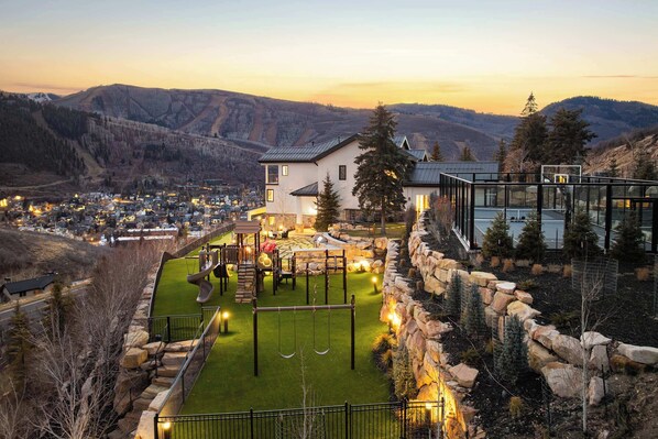 A residential area with a house, a playground, and a view of a town and mountains in the background. - Luxe PC Estate | Chef’s Kitchen + Acreage + Views (Park City)