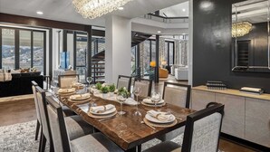 Ein Essbereich mit einem Holztisch, der für sechs Personen gedeckt ist, umgeben von Stühlen. Eine Kristallleuchter hängt darüber, und das Zimmer hat große Fenster mit Blick ins Freie.