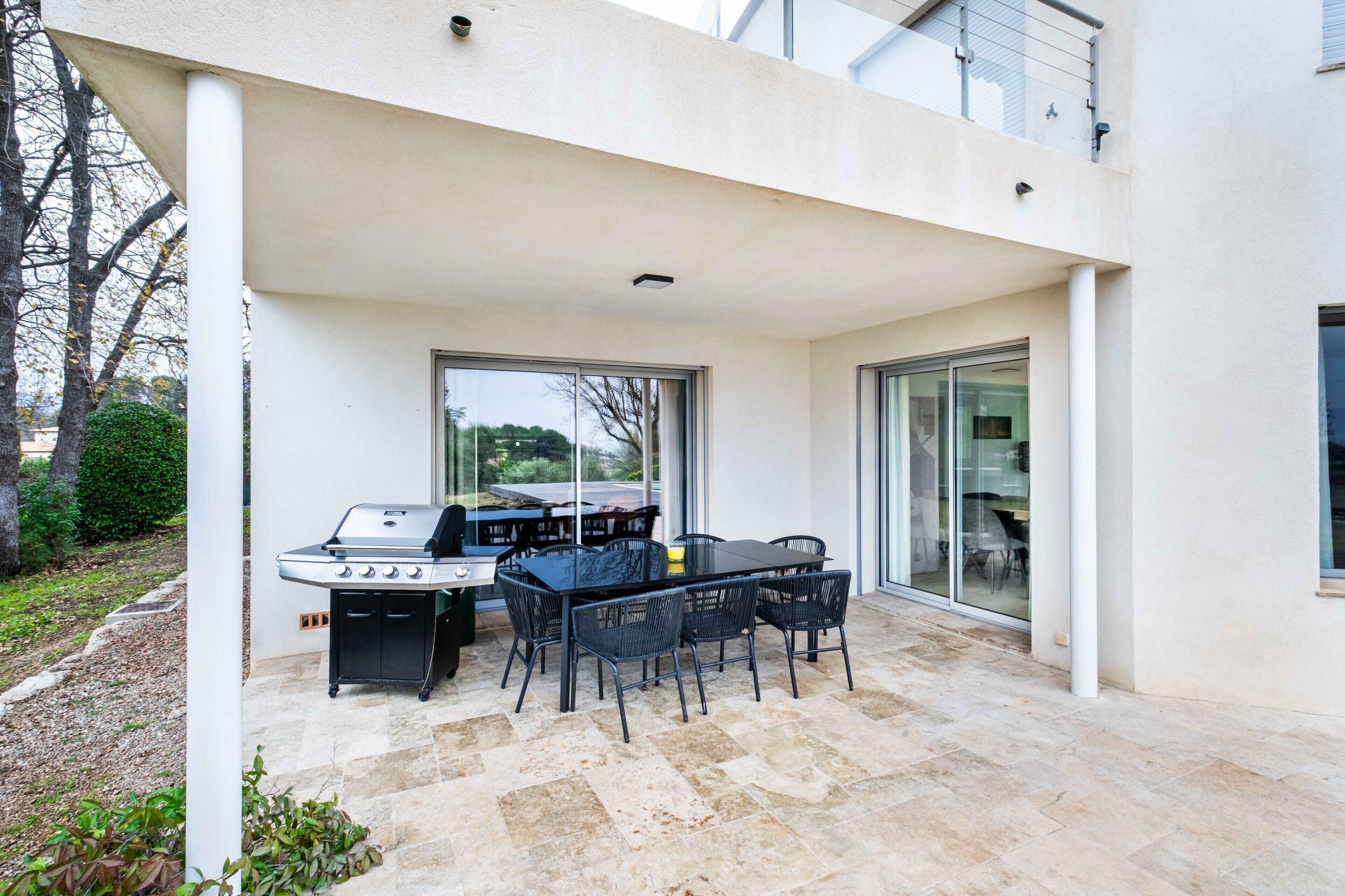 Un patio exterior con una parrilla, una mesa de comedor y sillas, al lado de un edificio con ventanas amplias y un balcón.