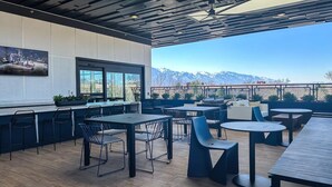 Eine Terrasse im Freien mit Tischen, Stühlen, einem Wandfernseher und Blick auf Berge.