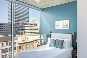 Een hotelkamer met een groot raam met uitzicht op de stad, een bed met witte lakens en gestreepte kussens, een nachtkastje met een lamp en een ingelijst kunstwerk aan de muur.
