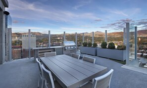 Een buitenpatio met een eettafel en stoelen, een barbecue en een uitzicht op een stadsbeeld met bergen op de achtergrond.