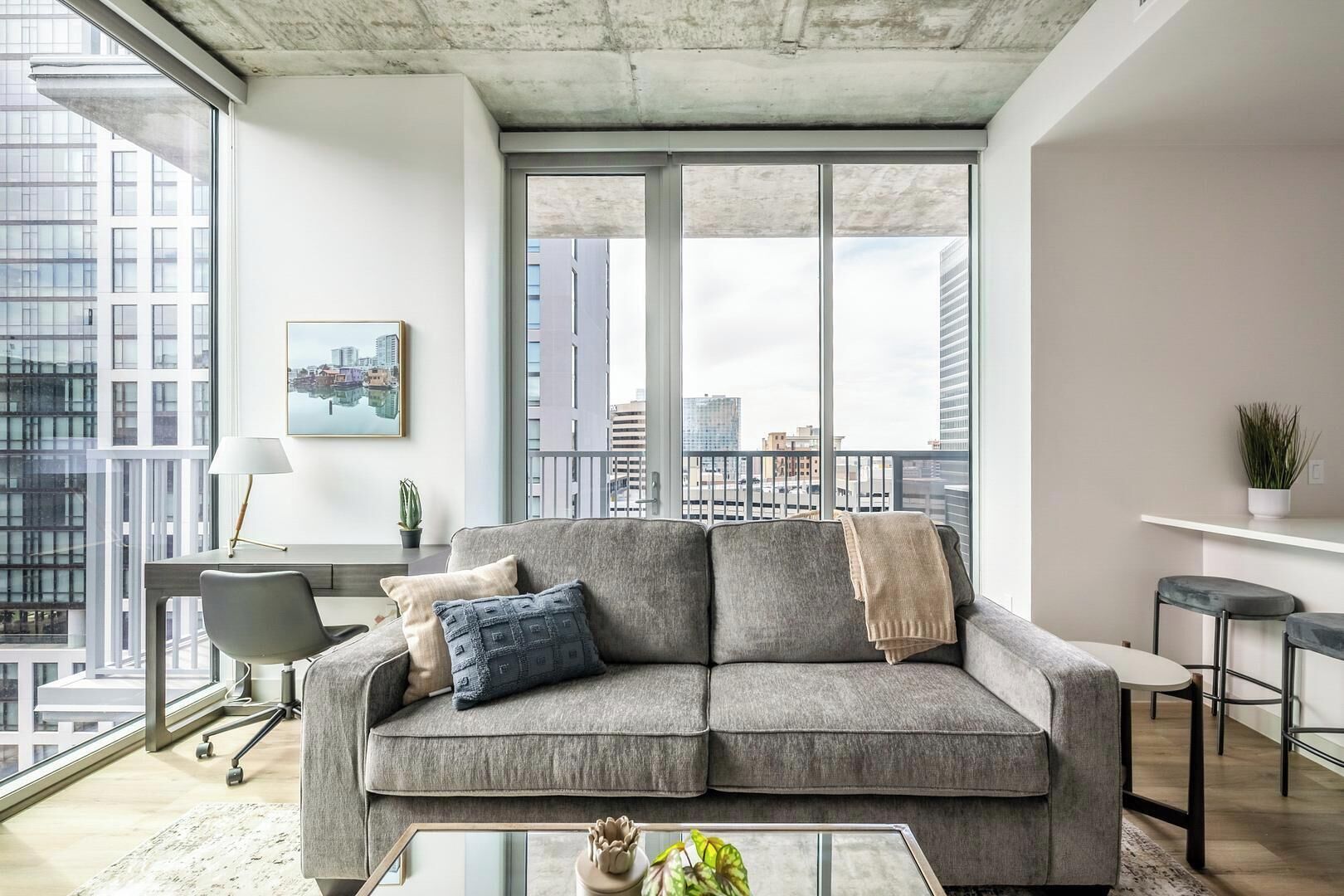 A modern living room with a grey sofa, a glass coffee table, and a city view through large windows.