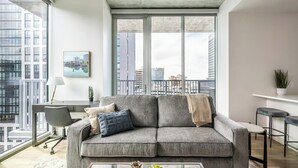 Ein modernes Wohnzimmer mit grauem Sofa, einem Glastisch und Blick auf die Stadt durch große Fenster.