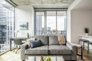 Ein modernes Wohnzimmer mit grauem Sofa, einem Glastisch und Blick auf die Stadt durch große Fenster.