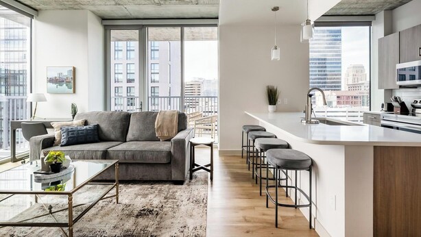 Eine moderne Wohnung mit einem grauen Sofa, einem kleinen Esstisch und einer Küche mit Bar.