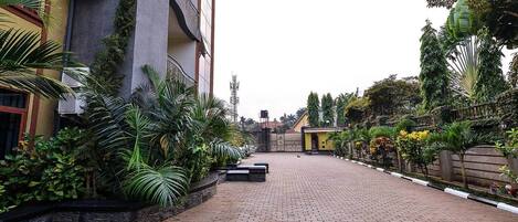 A paved courtyard with a building on one side and a variety of plants and trees on the other.