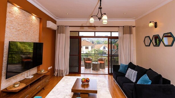 Una sala de estar con televisión de pantalla plana, una mesa de centro de madera con fruta, un sofá negro con cojines azules y un balcón con mobiliario de mimbre.