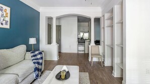 Una sala de estar con sofá, mesa de centro y lámpara. Se ve un área de cocina a través de un umbral abierto.