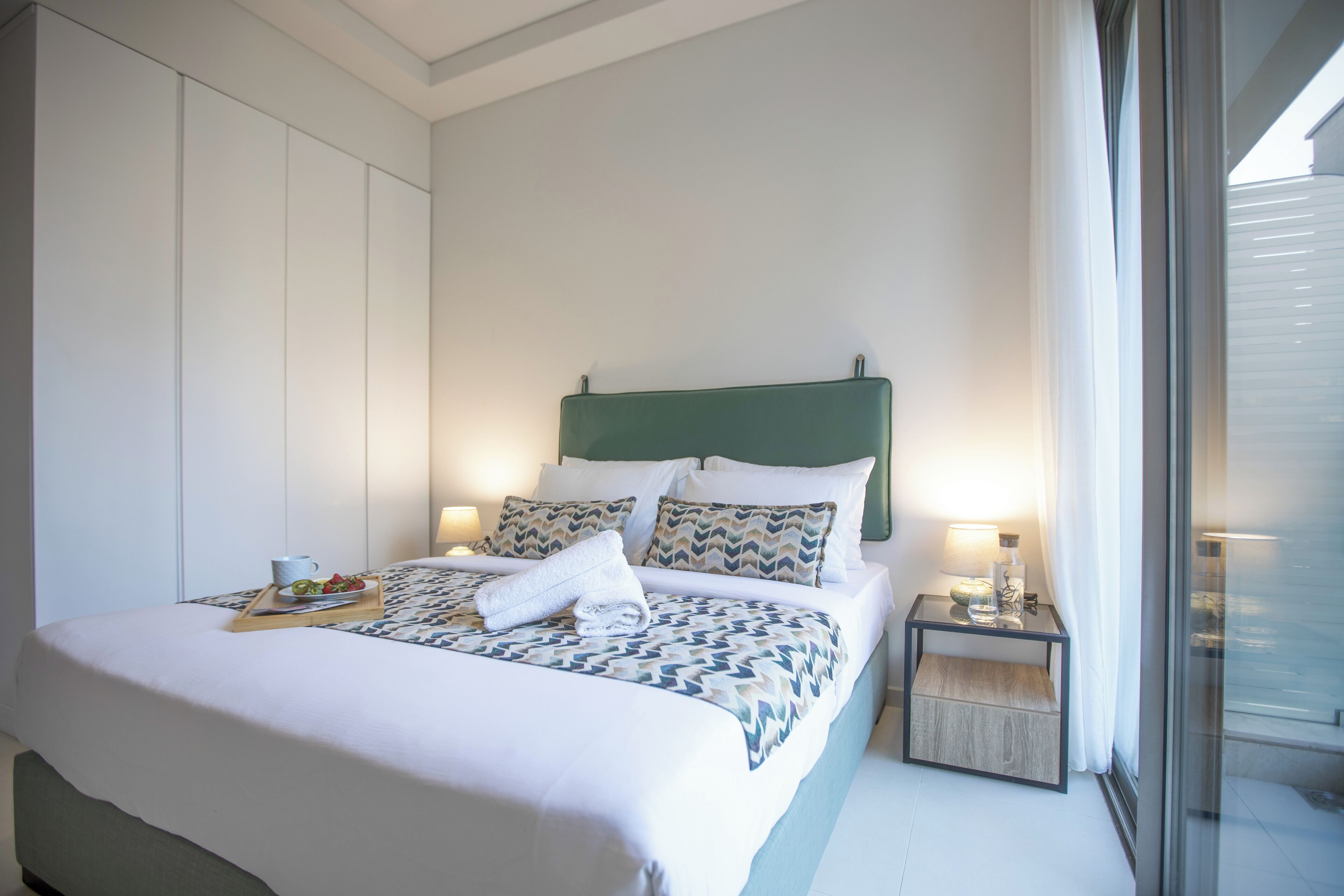 A neatly made bed with a patterned bedspread, a tray with a cup and food, and a bedside table with a lamp.