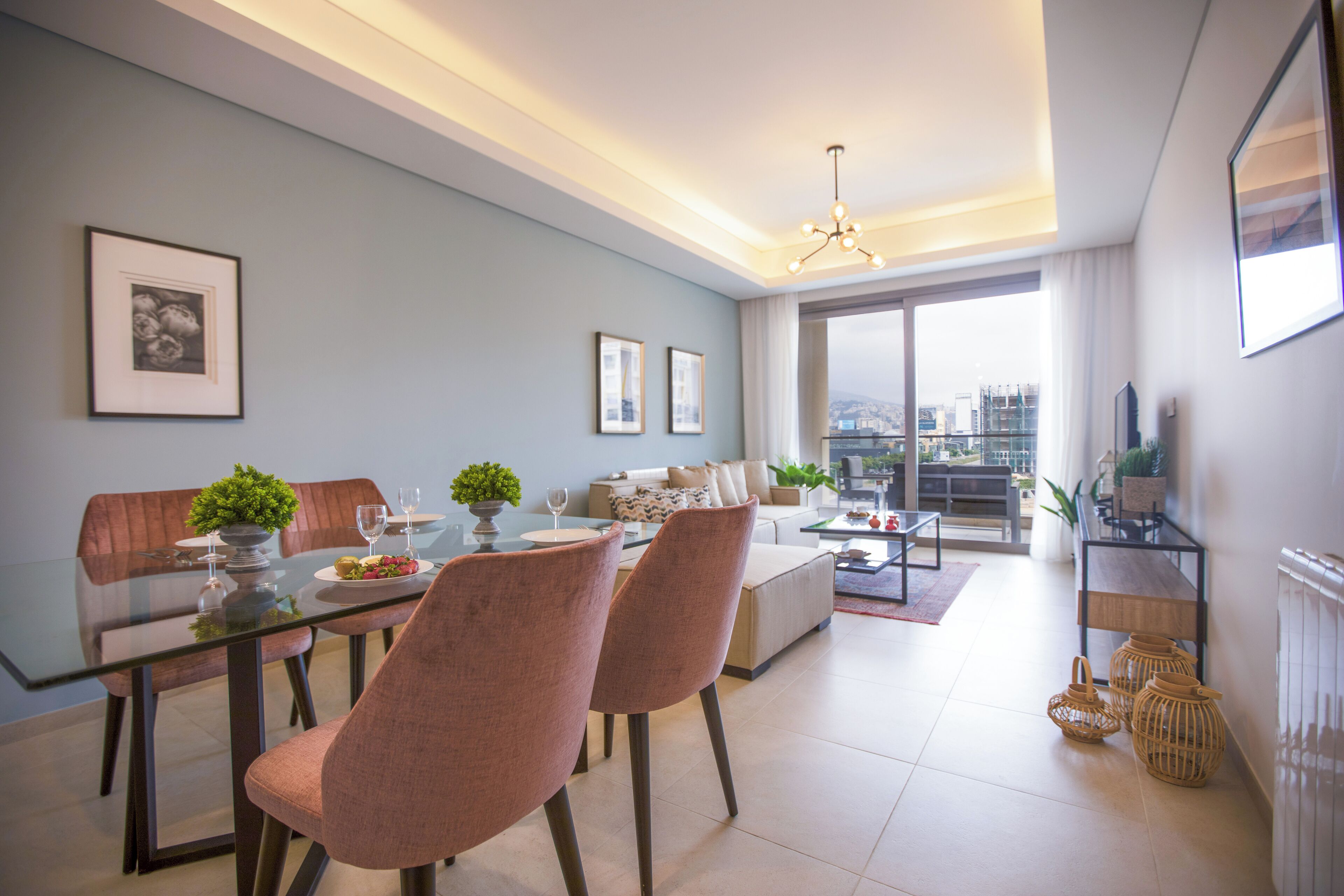 A modern living room with a glass dining table, two cushioned chairs, a sofa, and a coffee table. There are framed pictures on the wall and a large window with a city view.