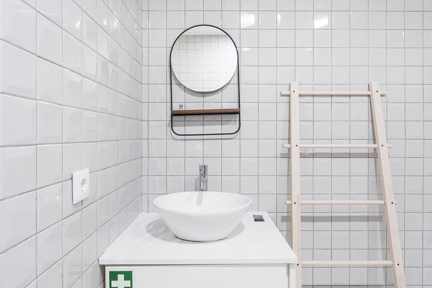 Ein Badezimmer mit weißen Fliesen, einem weißen Waschbecken und einem Spiegel darüber sowie einer Holzleiter.