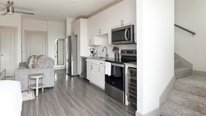 Una cocina moderna con electrodomésticos de acero inoxidable, un horno empotrado y un microondas. Hay un sofá gris y un taburete pequeño en la zona de estar contigua.