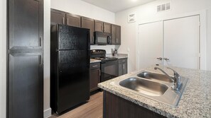 A modern kitchen with dark wood cabinets, a black refrigerator, a microwave, an oven, and a granite countertop with a double sink. - Landing | Chic 1BD, Pool, Clubhouse (San Antonio)