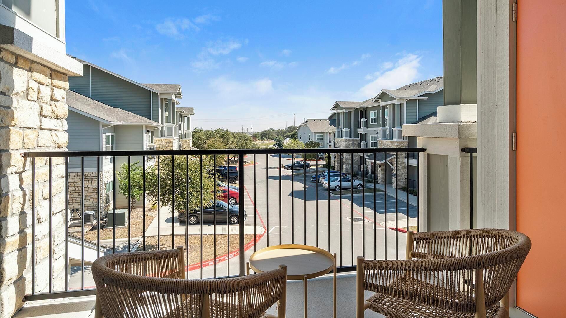 A balcony with wicker chairs and a small table, overlooking a parking lot and adjacent buildings.