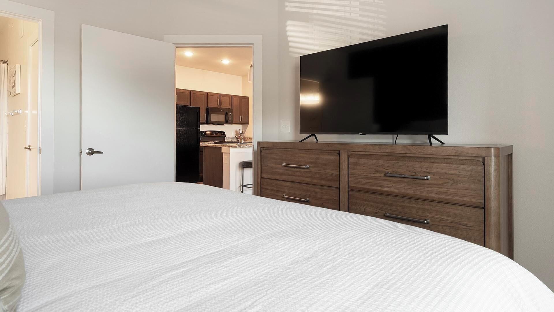 A bedroom with a large bed, a TV mounted on a wooden dresser, and a view into the kitchen.