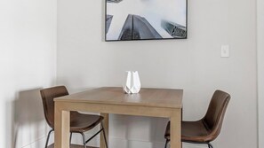A modern dining area with a wooden table and two leather chairs, a white vase on the table, and a framed cityscape picture on the wall. - Landing | Incredible 1BD, Pool, Clubhouse (San Antonio)