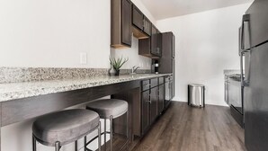 A modern kitchen with dark wood cabinets, a granite countertop, and a stainless steel refrigerator. - Landing | Incredible Studio, Clubhouse, Gym (San Antonio)