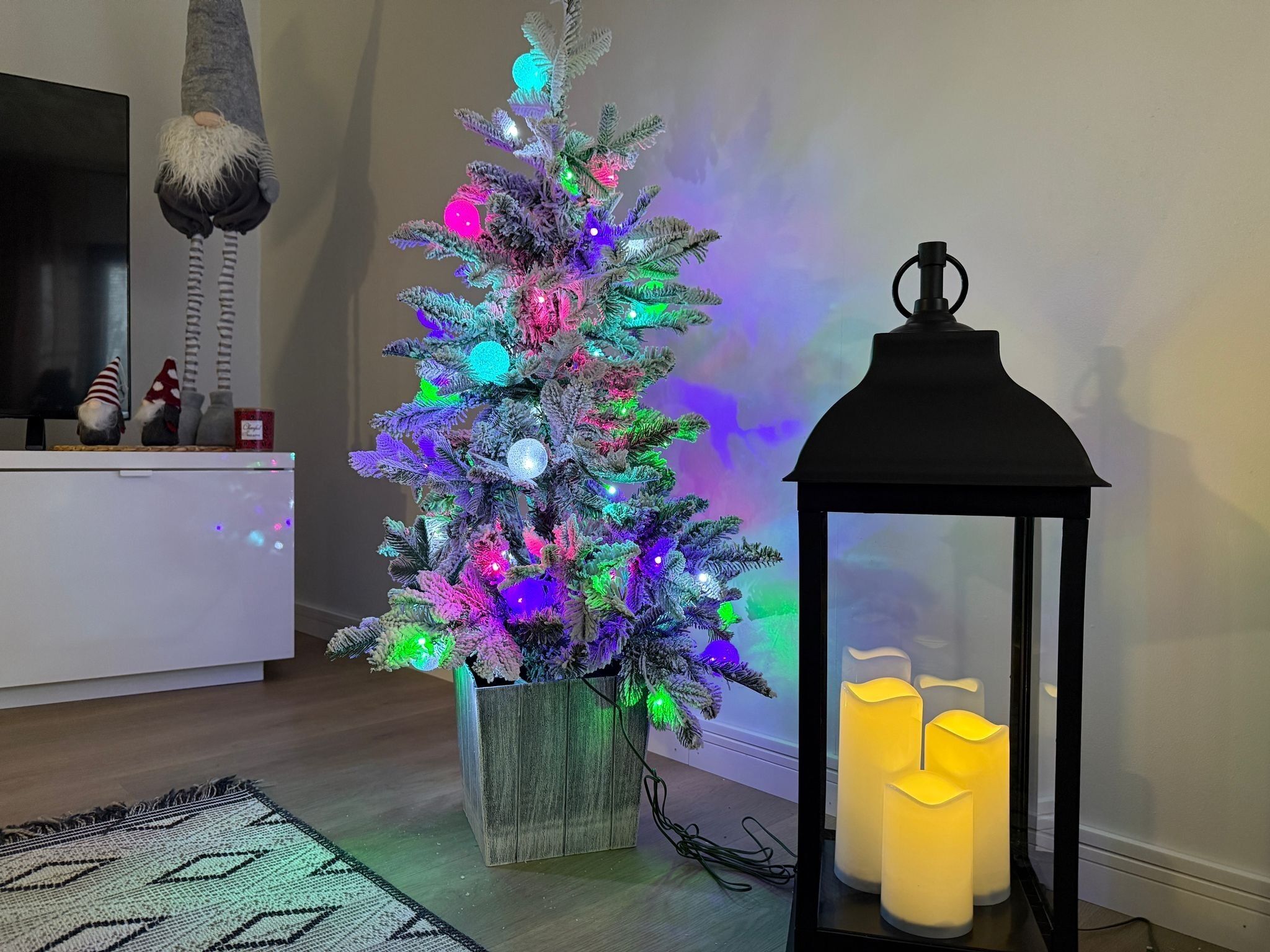 Ein Weihnachtsbaum mit bunten Lichtern, ein schwarzer Laternenleuchter mit Kerzen und ein gemustertes Teppich in einem Raum mit einem weißen Schrank und einem Fernseher.