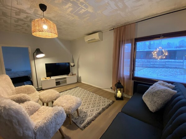 A modern living room with a flat-screen TV, fluffy chairs, a sofa, a patterned rug, and a ceiling light fixture.