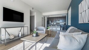 A modern living room with a flat-screen TV, a grey sofa, a coffee table, and a kitchen in the background. - Landing | Incredible 1BD, Tennis Court, Gym (Fort Myers)