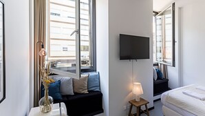A modern hotel room with a large window, a flat-screen TV mounted on the wall, a bedside table with a lamp, and a bed with white bedding. - Regras Apartments & Garden A9 by Benefit Collection (Porto)