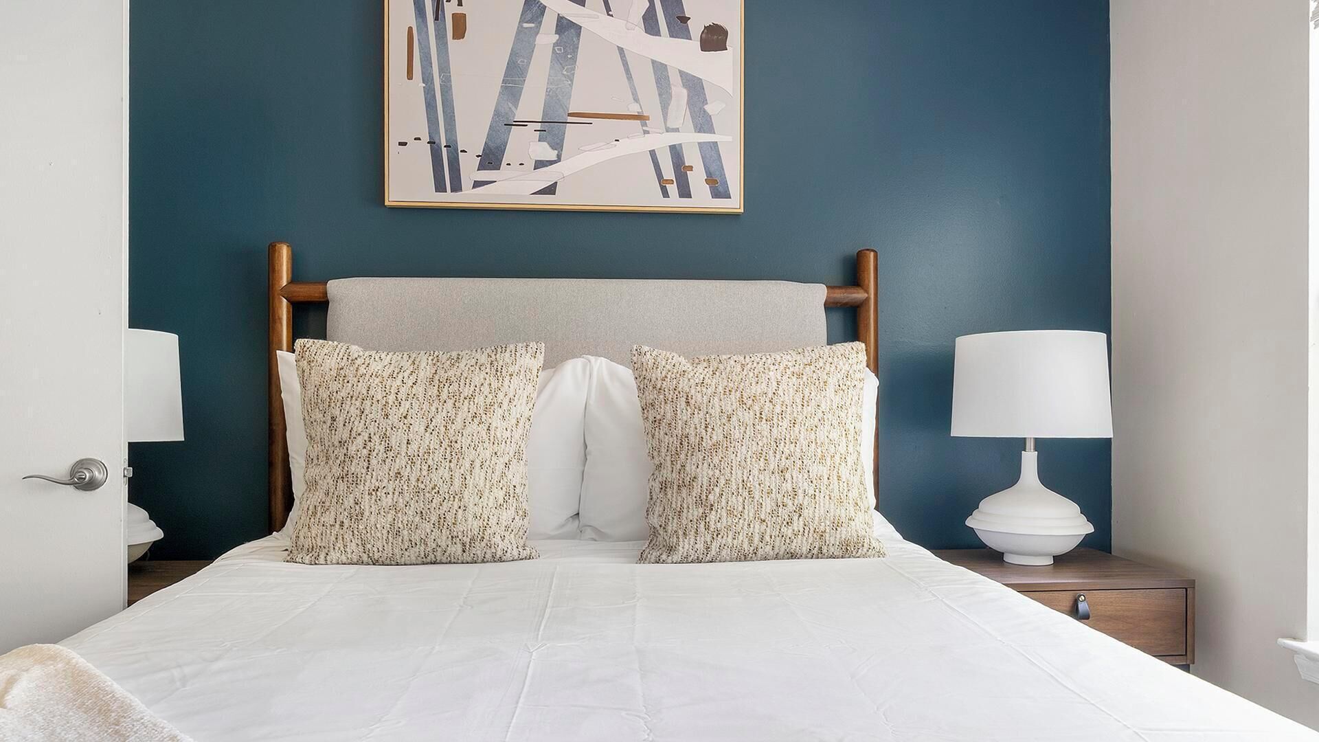 A bedroom with a bed, two pillows, a nightstand, a lamp, and a painting on the wall.