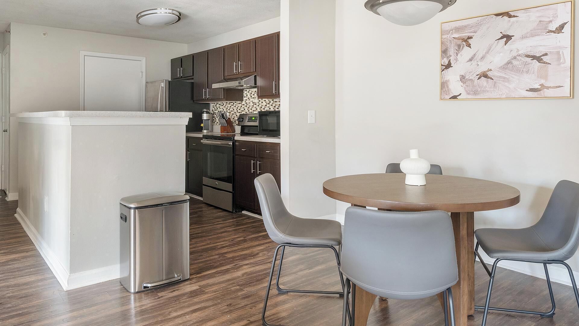 A compact kitchen with dark cabinets, a stainless steel refrigerator, and a microwave. There is a small trash bin and a round table with two chairs.