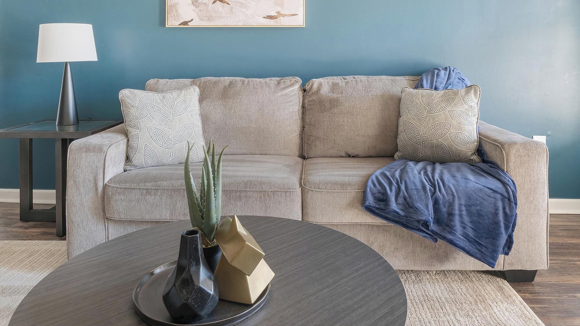 A living room with a beige sofa, patterned pillows, a blue throw blanket, a coffee table with a vase and a plant, and a lamp on a side table.