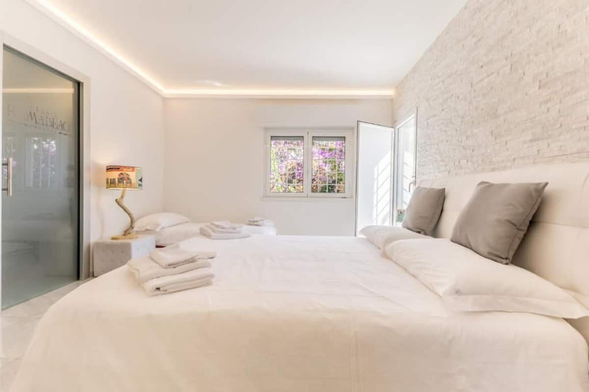 Quarto com cama grande, mesa de cabeceira, luminária e janela com vista para flores.
