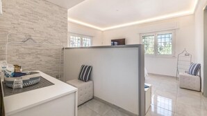 A modern, well-lit room with a white brick wall, a cushioned bench, a small table with a basket, and a view of the outdoors through large windows. - RELAIS ROMA BEACH (Fiumicino)