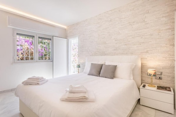 A modern bedroom with a large bed, white bedding, a brick accent wall, and a bedside table with a lamp. - RELAIS ROMA BEACH (Fiumicino)