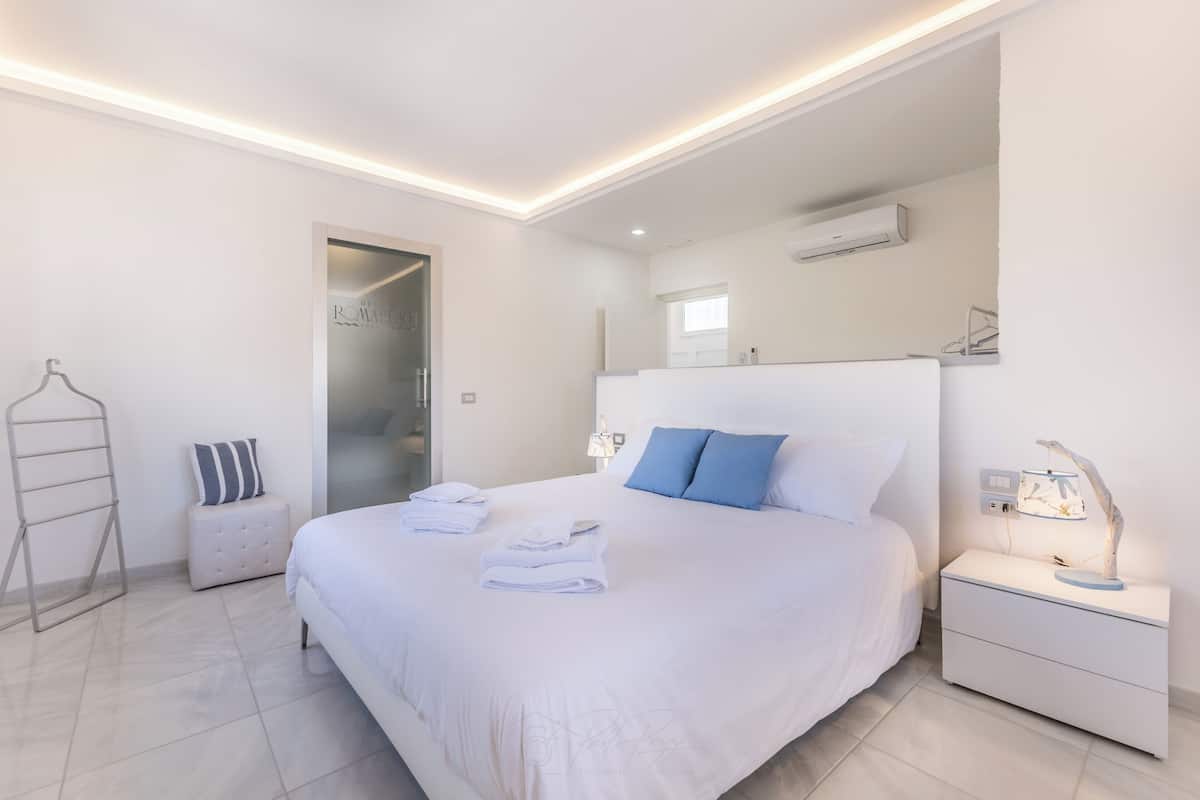 Quarto de hotel moderno com uma cama grande, uma mesa de cabeceira com abajur, um cabideiro e um banquinho pequeno.