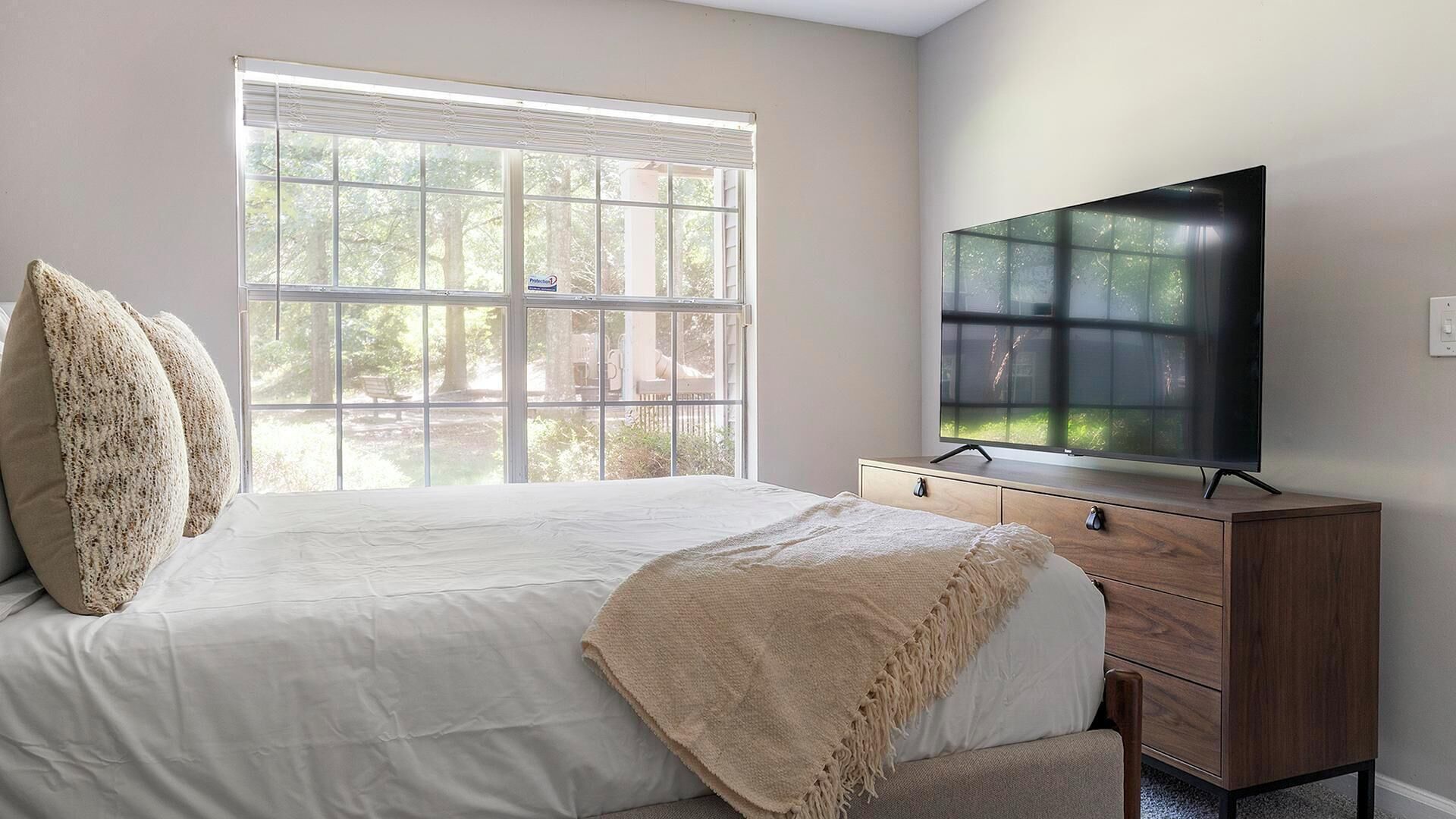 Een slaapkamer met een groot raam, een bed, een tv op een houten dressoir en een kast.