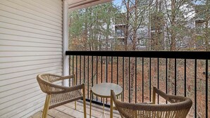 Een balkon met rieten stoelen en een klein rond tafeltje, met uitzicht op een bebost gebied met bomen en een gebouw op de achtergrond.