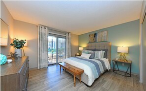 A bedroom with a bed, bedside tables, a bench, a desk, and a view through a sliding door to an outdoor area. - Landing | Modern 1BD, Pool, Clubhouse (Huntsville)