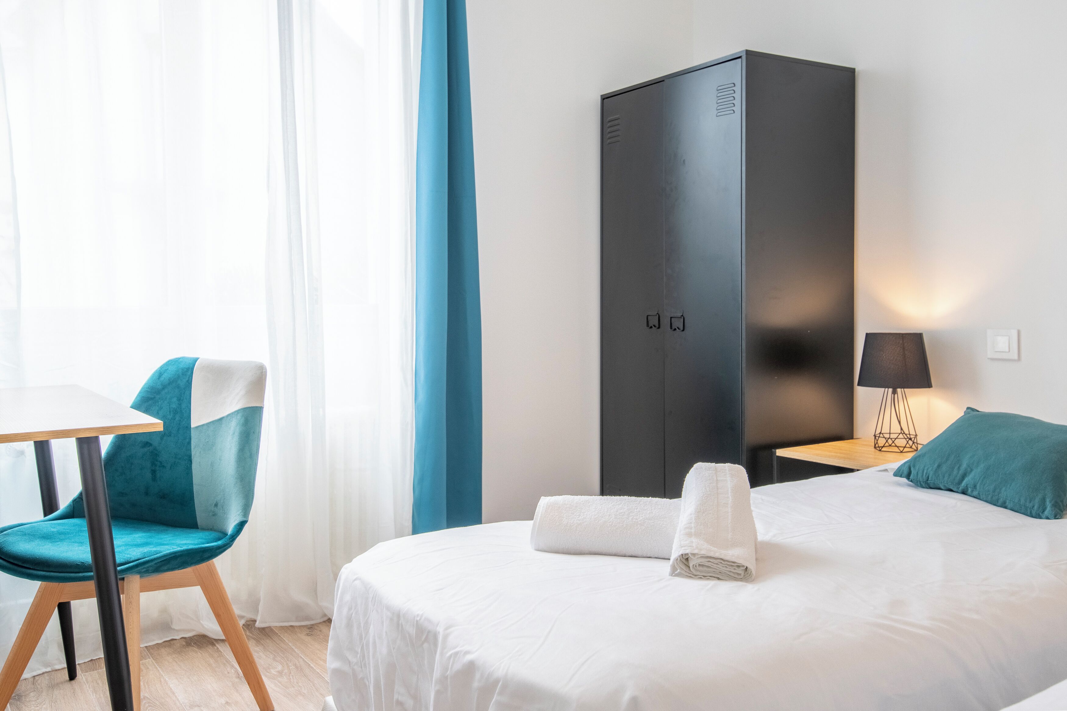 A hotel room with a bed, a chair, a desk, a wardrobe, and a lamp.