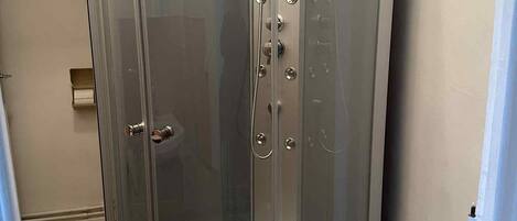 A glass-enclosed shower stall with a handheld showerhead, a control panel, and a drain.