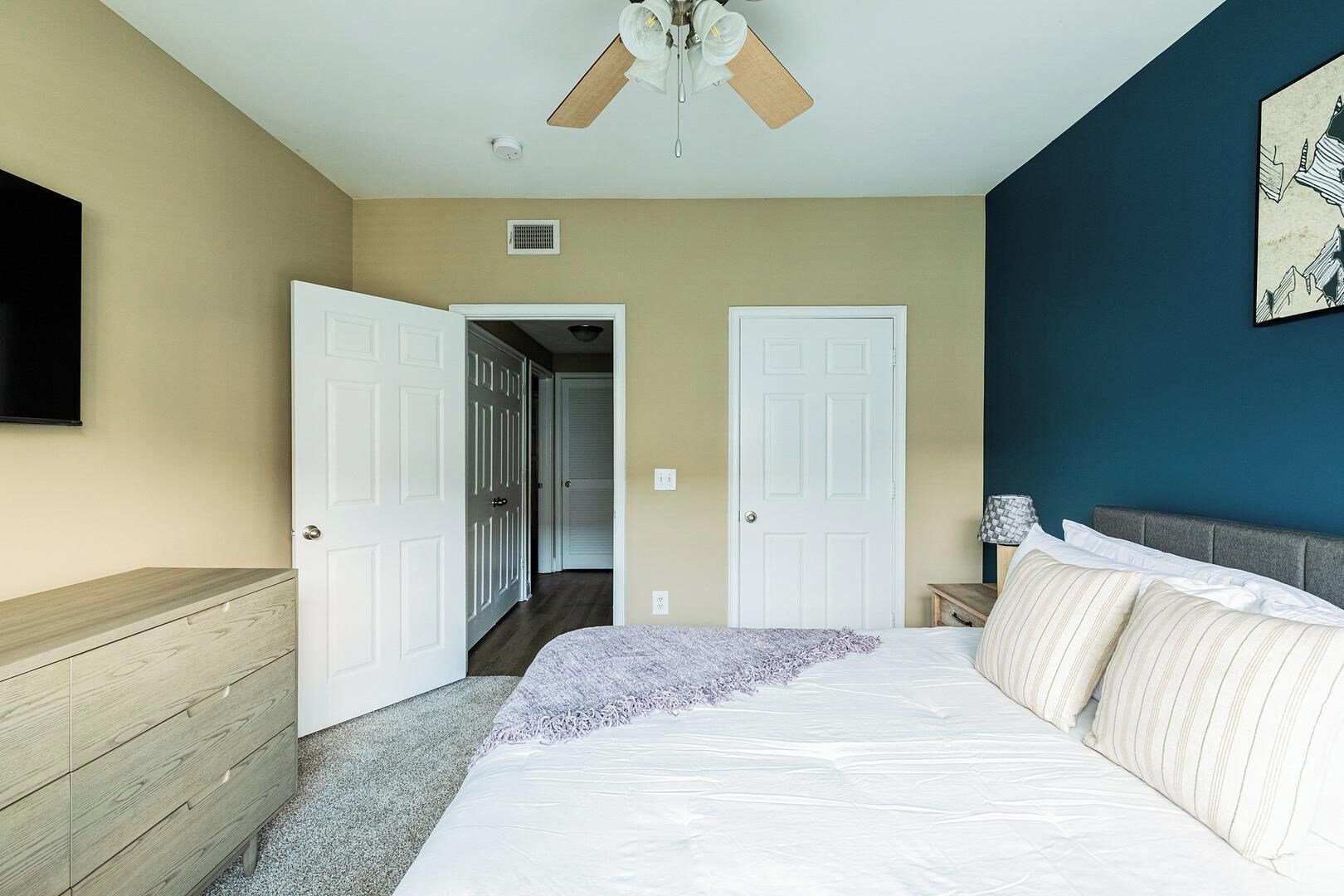 A bedroom with a bed, a ceiling fan, a TV mounted on the wall, and a hallway leading to another room.