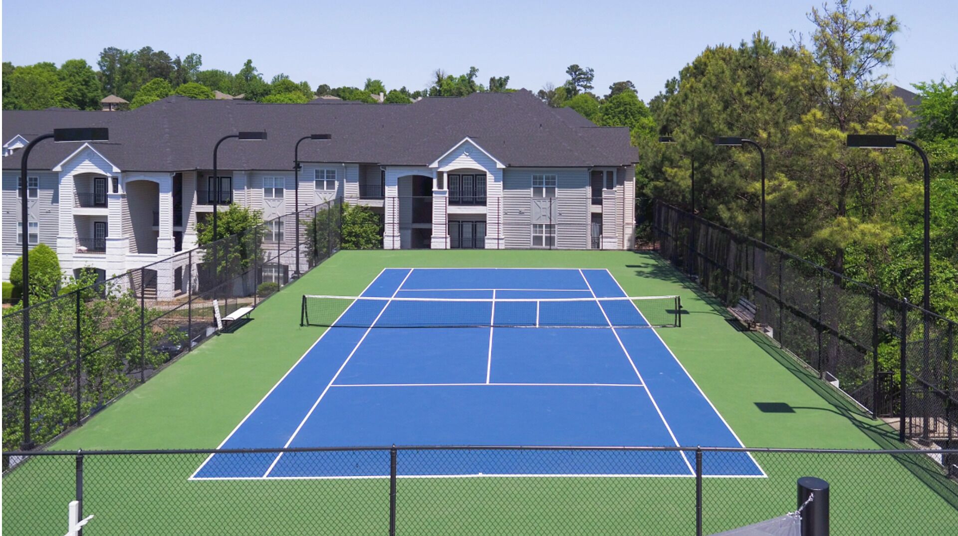 Un court de tennis avec un filet, entouré d’une clôture, et situé à côté d’un bâtiment dont le toit est foncé et les murs blancs.