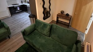 A living room with a green sofa, a wooden side table, and a kitchen in the background. - Stunning 2 Bed 2 Bath City Centre Apartment Liverpool (Liverpool)