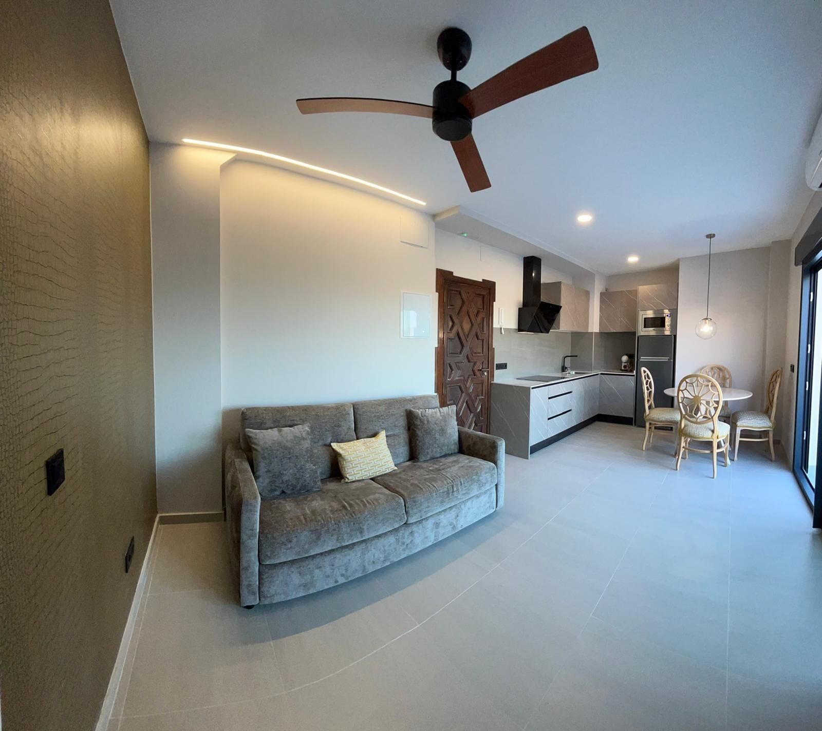 A modern living room with a grey sofa, a ceiling fan, and a kitchen area with a dining table and chairs.