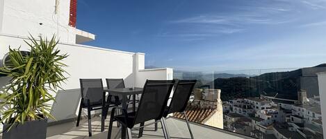 A rooftop terrace with a table and chairs, a potted plant, and a view of a town and hills.