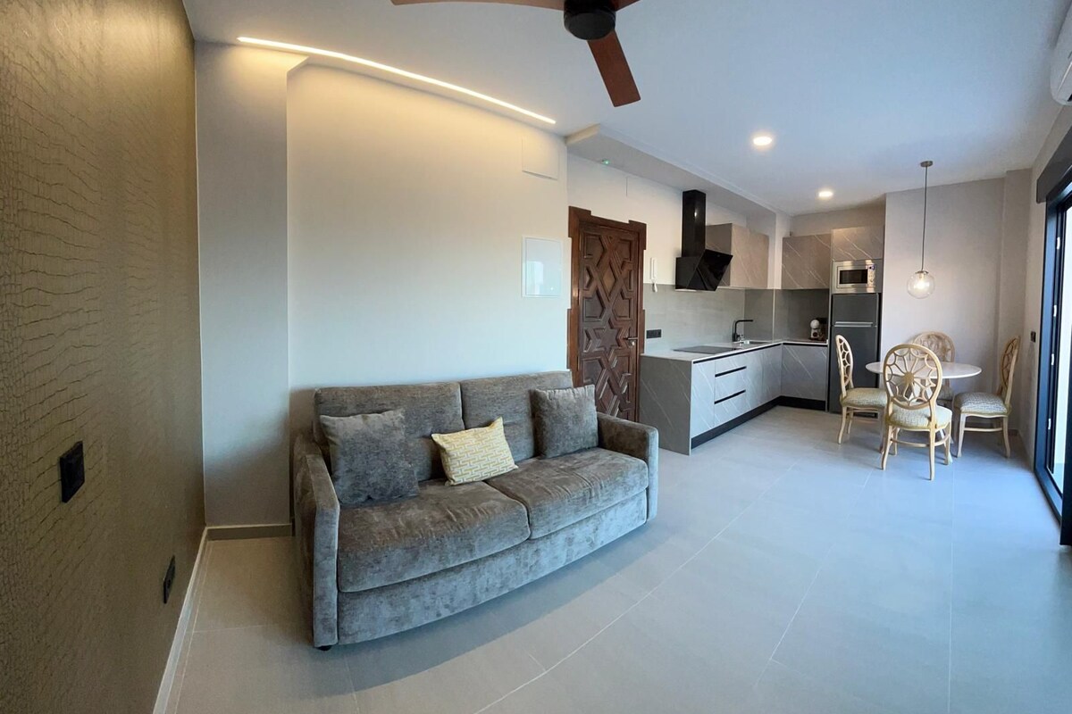 A modern living room with a grey sofa, a ceiling fan, and a kitchen area with a dining table and chairs.