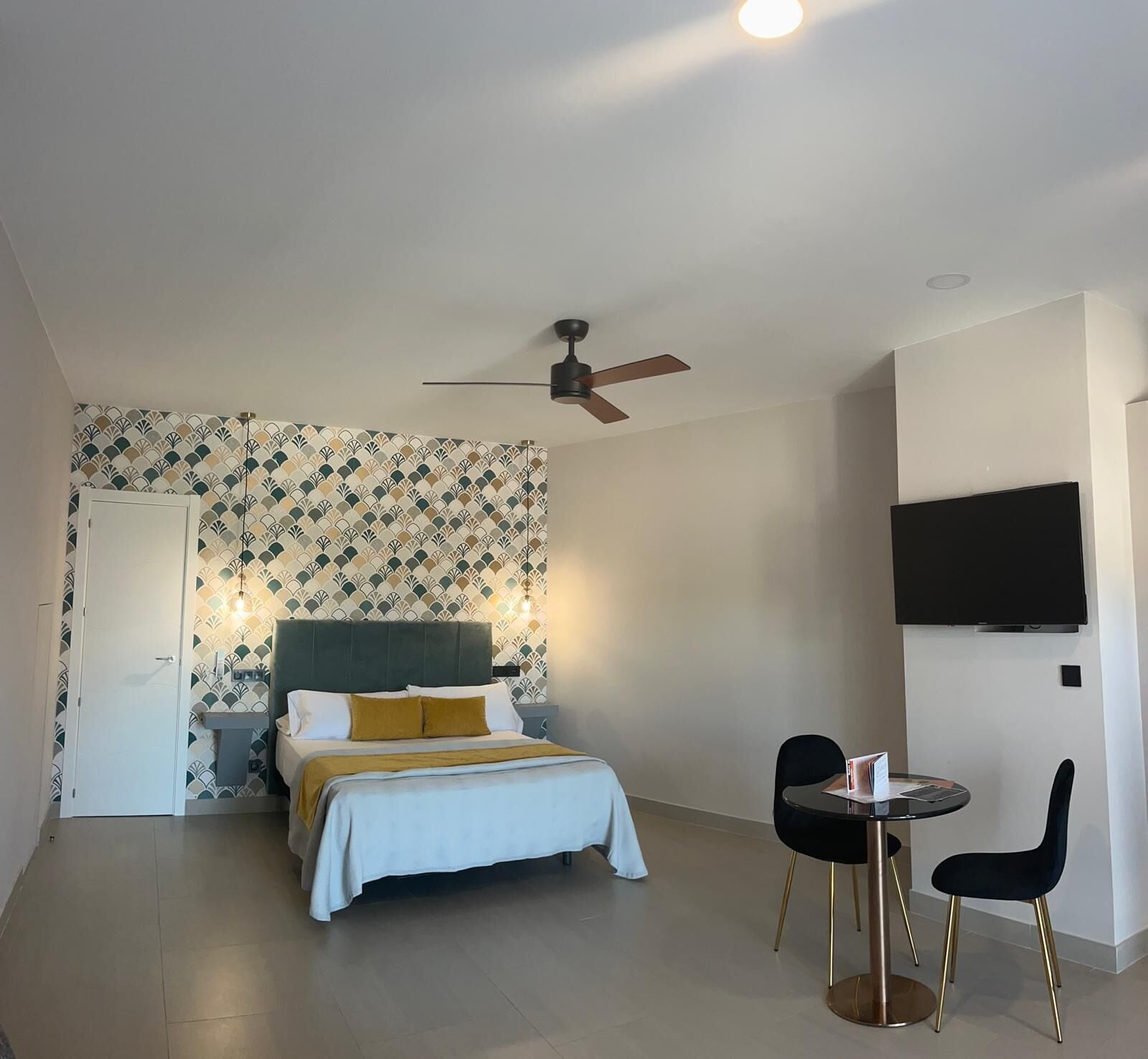 Un dormitorio con cama, escritorio con silla, ventilador de techo y televisor montado en la pared.